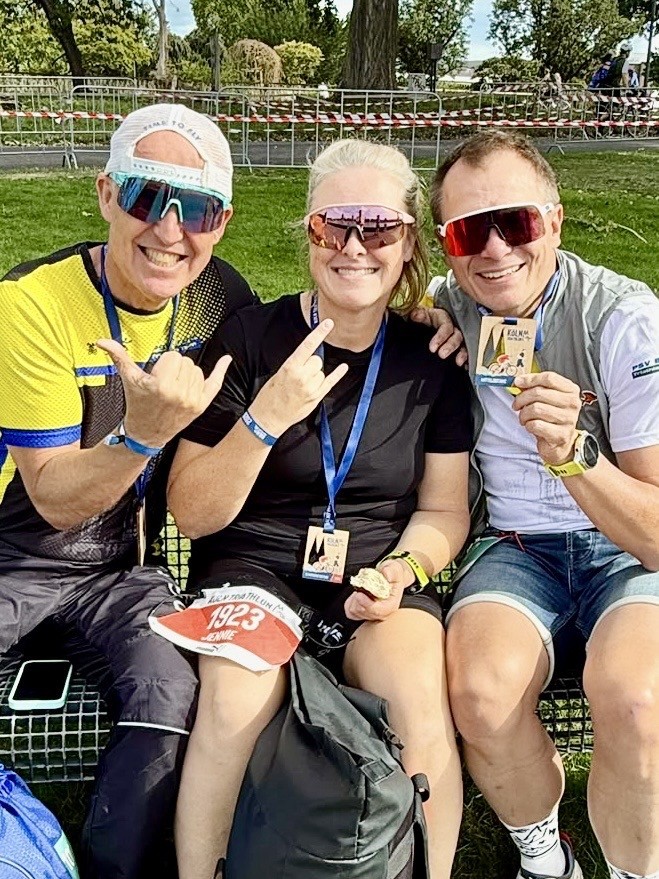 Drei Personen mit Finisher-Medaillen und Urkunden des Köln Triathlon sitzen auf einer Bank, in der Mitte Jennie Schaaf vom PSV Brühl
