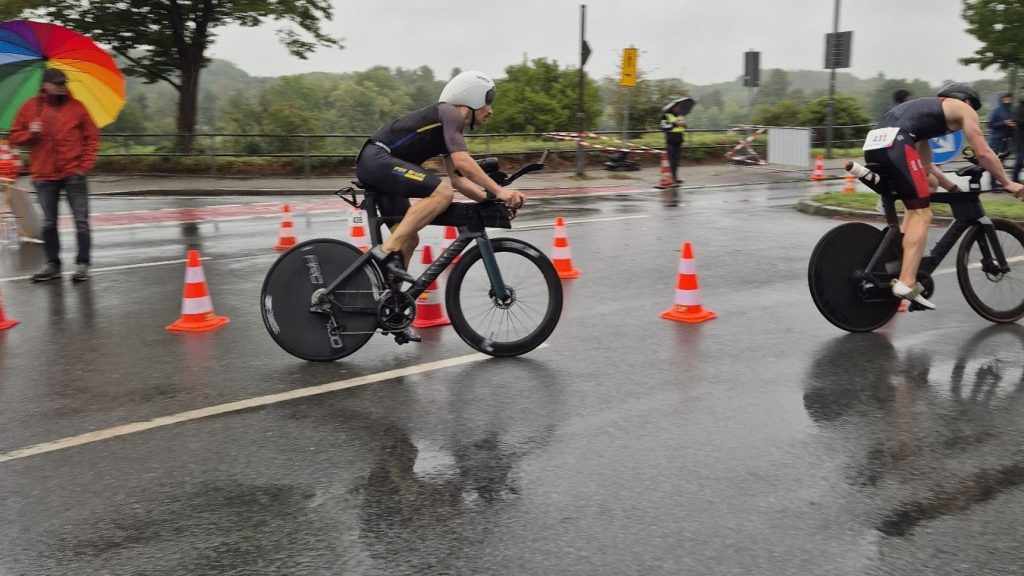 Zwei Triathleten auf Zeitfahrrädern bei strömendem Regen auf der Radstrecke des Oberliga-Finales in Ratingen, im Vordergrund ein Zuschauer mit Regenbogenschirm