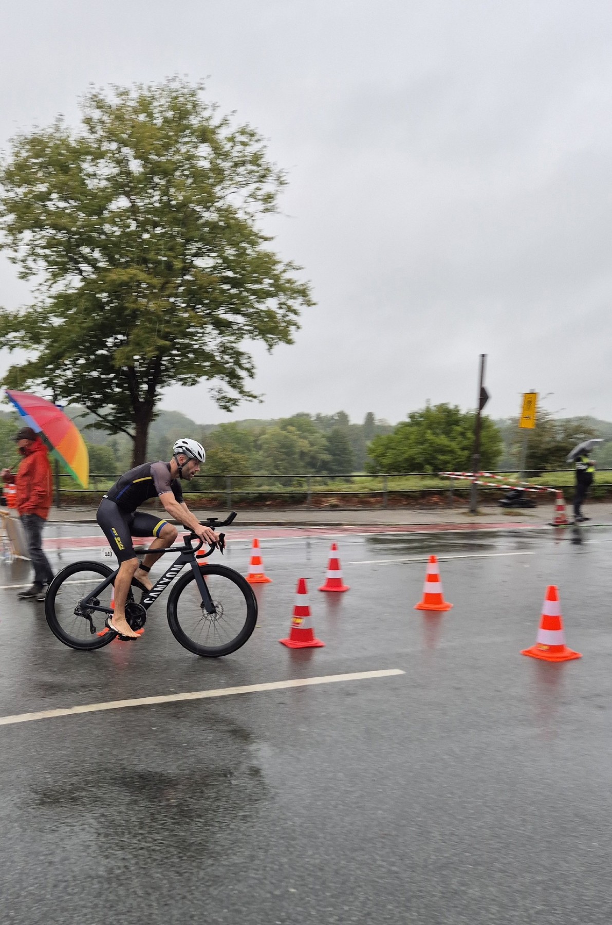 Athlet des PSV Brühl Triathlon Team im Trikot auf der nassen Radstrecke in Ratingen, im Hintergrund Zuschauer mit Regenbogenschirm